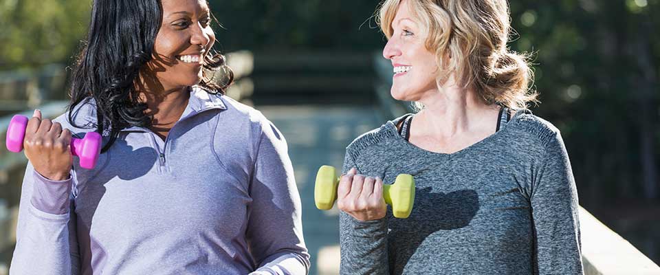 Two energetic women smile at each other in the park while lifting small weights.