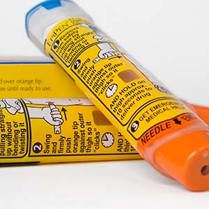 An EpiPen with an orange tip lies across a second EpiPen, with the box in the background.