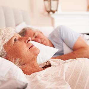 An elderly woman lies in bed and stares at the ceiling while her partner sleeps peacefully beside her.