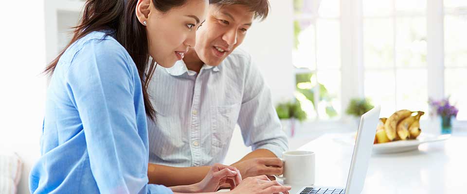 A husband and wife sit at a table in a room full of natural light, browsing their health plan on their computer.