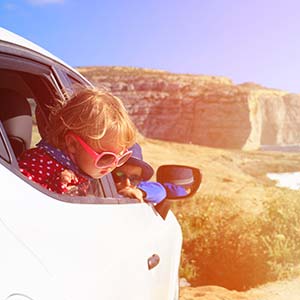 A little girl with oversized sunglasses peers out the window of the car as they drive on the coastline.