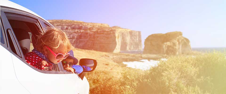 A little girl with oversized sunglasses peers out the window of the car as they drive on the coastline.