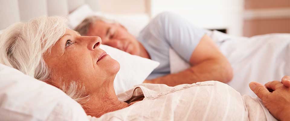 An elderly woman lies in bed and stares at the ceiling while her partner sleeps peacefully beside her.