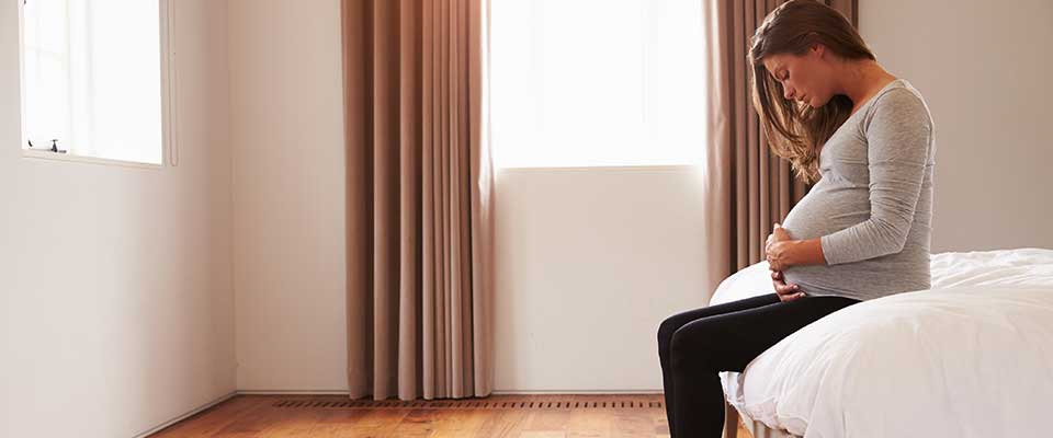 A young pregnant woman sits on the edge of her bed, looking down at her stomach as white light filters in through the window.