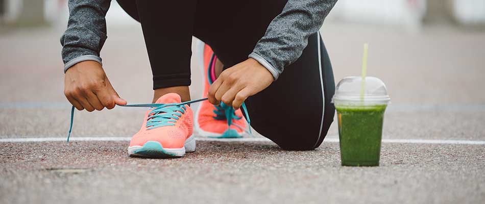 An athletic woman ties her shoelaces outside with a green juice propped next to her.