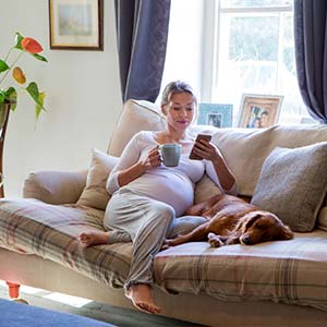 An expecting mother lounges on her couch, drinking caffeine during pregnancy. Her dog lies peacefully beside her.