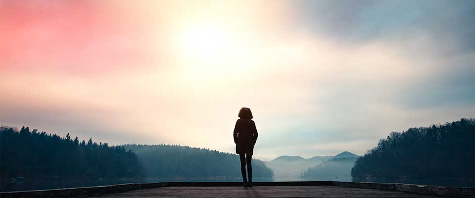 A woman stands at the edge of a serene lake. She is silhouetted by the sun. She keeps her hands in her pockets as she practices mindfulness meditation.