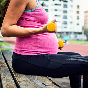 A woman is working out while pregnant. She sits on a bench alongside a city street, lifting small yellow weights.