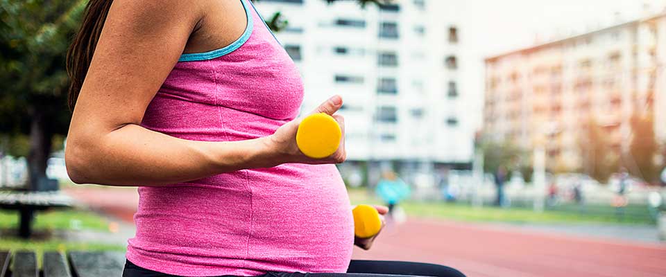 A woman works out while pregnant. She sits on a bench alongside a city street, lifting small yellow weights.