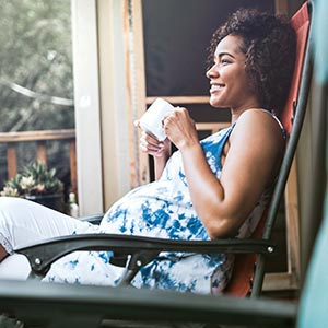 A pregnant woman relaxes on her back porch, smiling and sipping her tea.