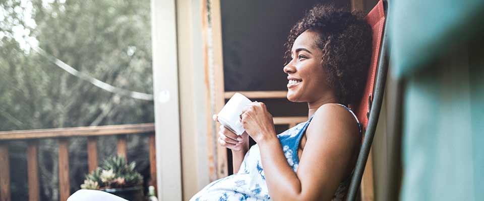 A pregnant woman relaxes on her back porch, smiling and sipping her tea.