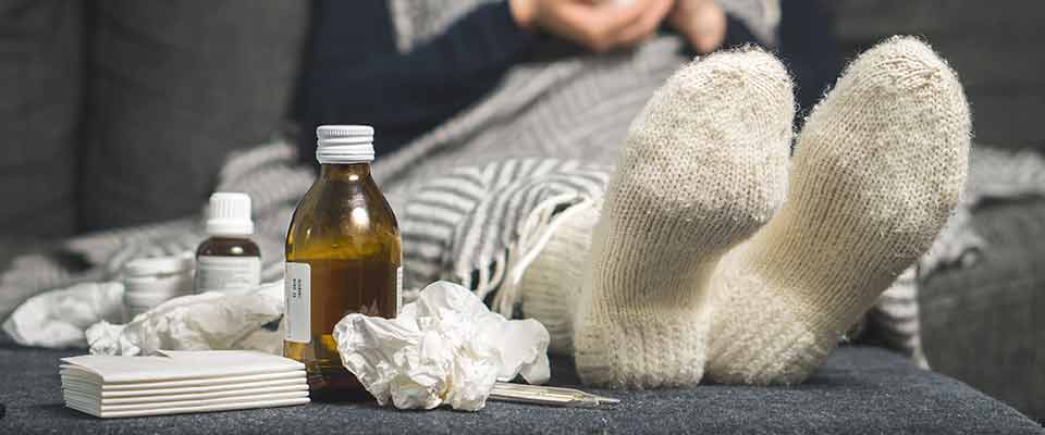 Kleenex, medicine, and a pair of feet lie near the edge of a couch, while their owner lies propped up, sneezing and sniffling.