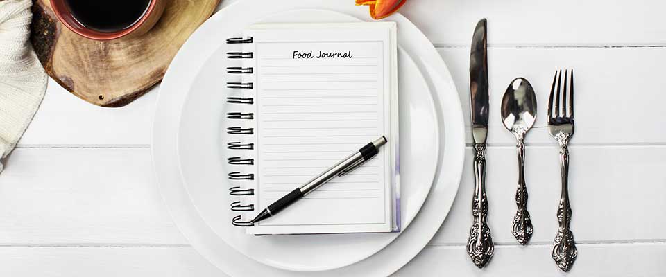 A pen and journal sit on a fancy white table between a set of silverware and a cup of coffee.