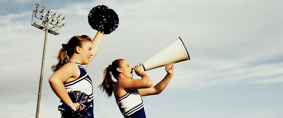 Two young cheerleaders at a high school game show their support with pom poms and a microphone.