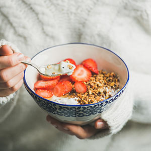 A woman in a white sweater spoons granola, yogurt, and strawberries from a bowl.