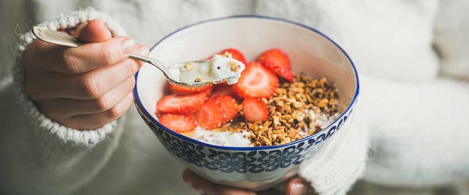 A woman in a white sweater spoons granola, yogurt, and strawberries from a bowl.