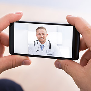 During a telehealth session, a pair of hands holds a phone displaying video of a smiling doctor in a white coat.