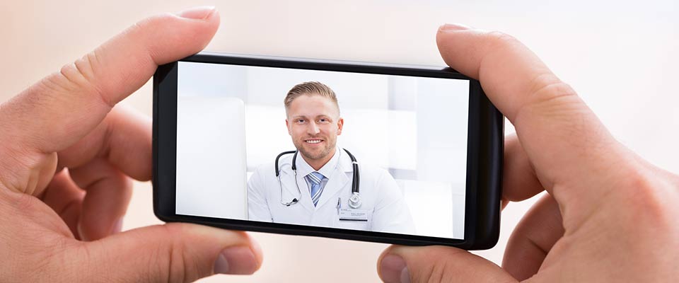 During a telehealth session, a pair of hands holds a phone displaying video of a smiling doctor in a white coat.