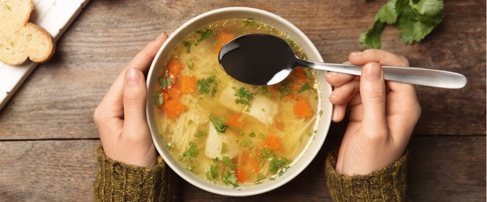 Hands holding a spoon and a bowl of chicken soup.